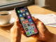 Main féminine tenant un smartphone avec applications d'économies, tasse de café et tickets de caisse sur une table en bois lumineuse